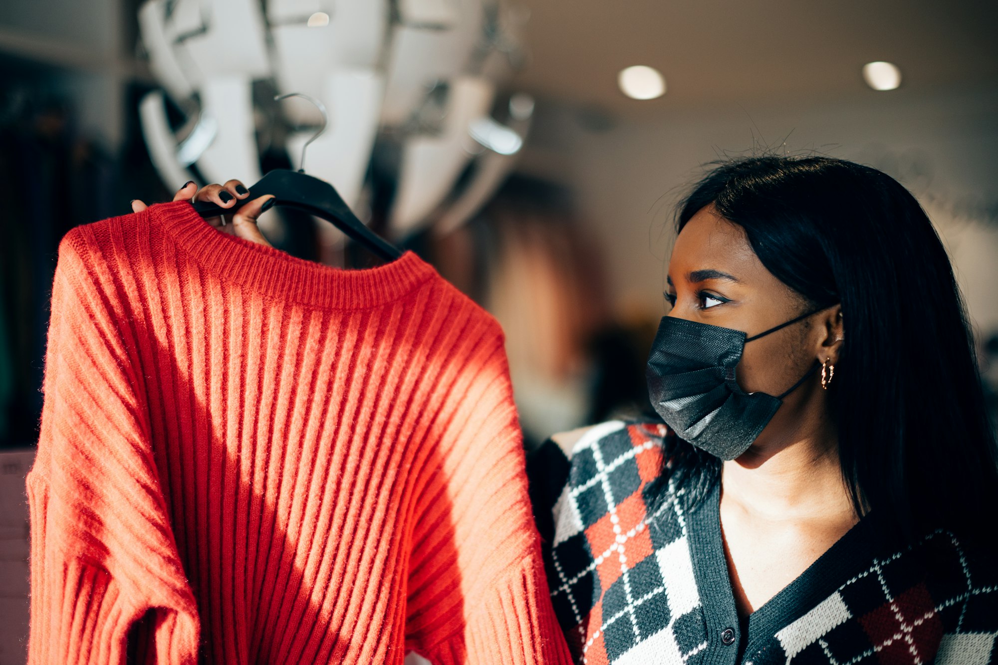 young woman with a black face mask standing in a fashion retail shop choosing a sweater